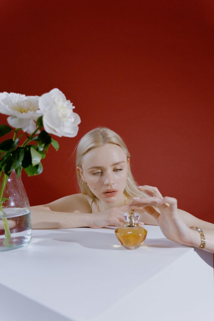 Elegant blonde woman reaching for perfume near white flowers.